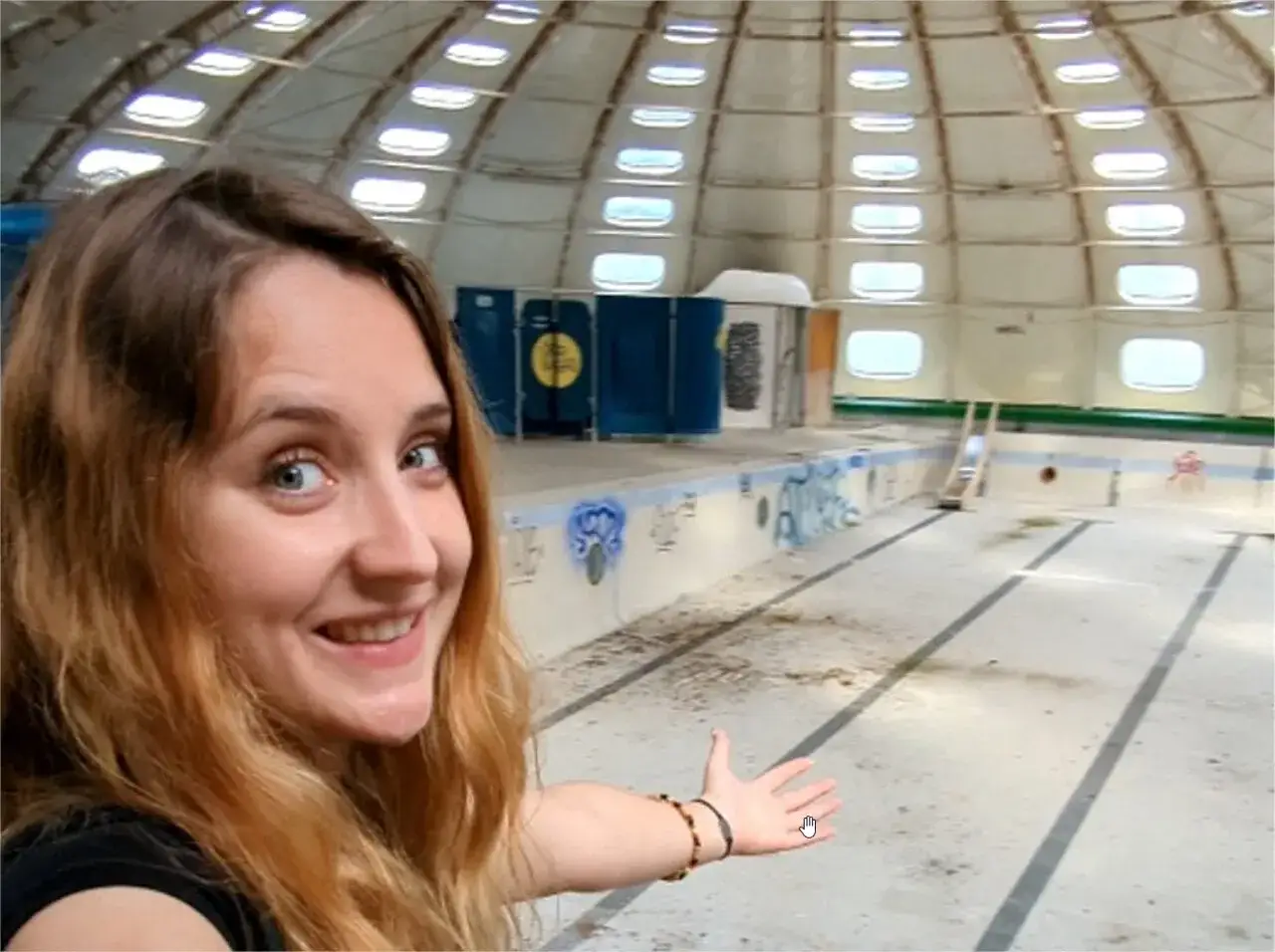 Une jeune femme souriante fait un geste vers une piscine tournesol abandonn&eacute;e, tagu&eacute;e et poussi&eacute;reuse, sous un d&ocirc;me lumineux.