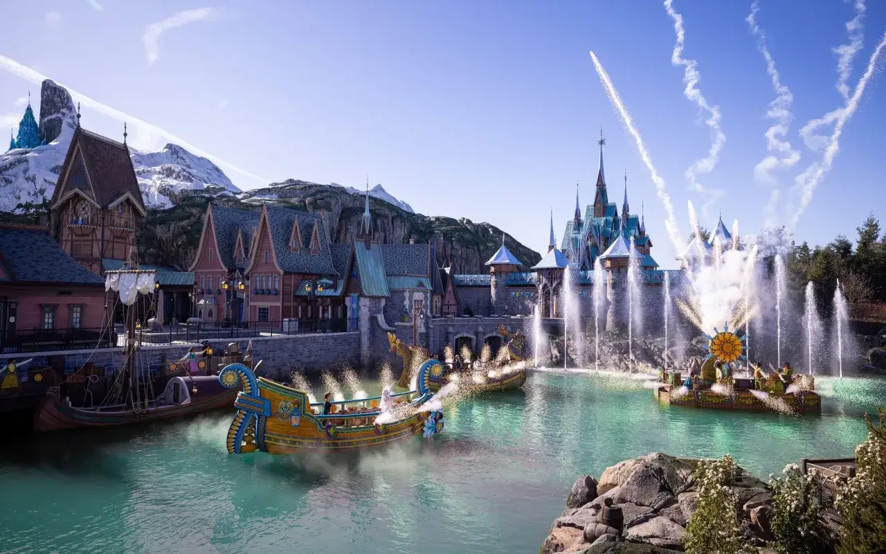 Lodzie płyną po wodzie, a w tle zamek i góry. Wspaniałe atrakcje Disneyland Paris z pokazem fontann i fajerwerków.