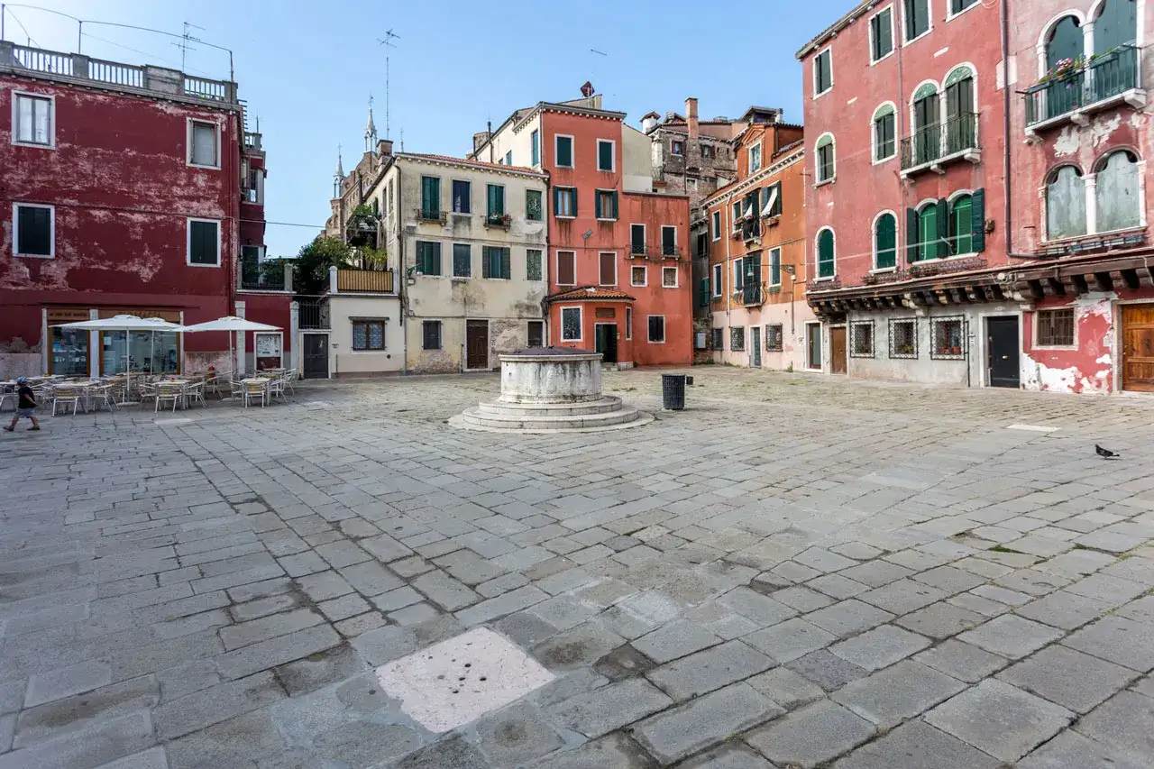 Una tranquilla piazza veneziana con un pozzo al centro, circondata da edifici colorati e tavolini all'aperto.