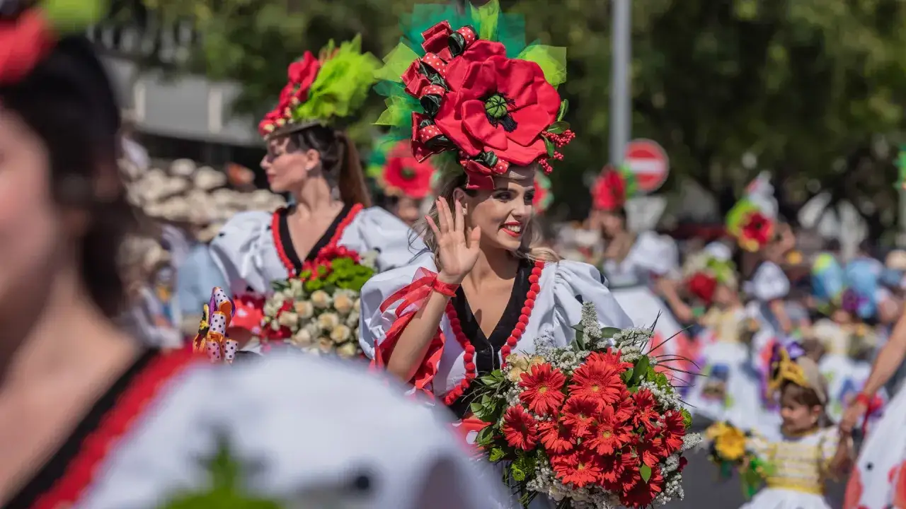 Uśmiechnięta kobieta w kwiecistej kreacji macha ręką podczas festiwalu kwiatów Madera. Wokół kolorowe stroje i bukiety.