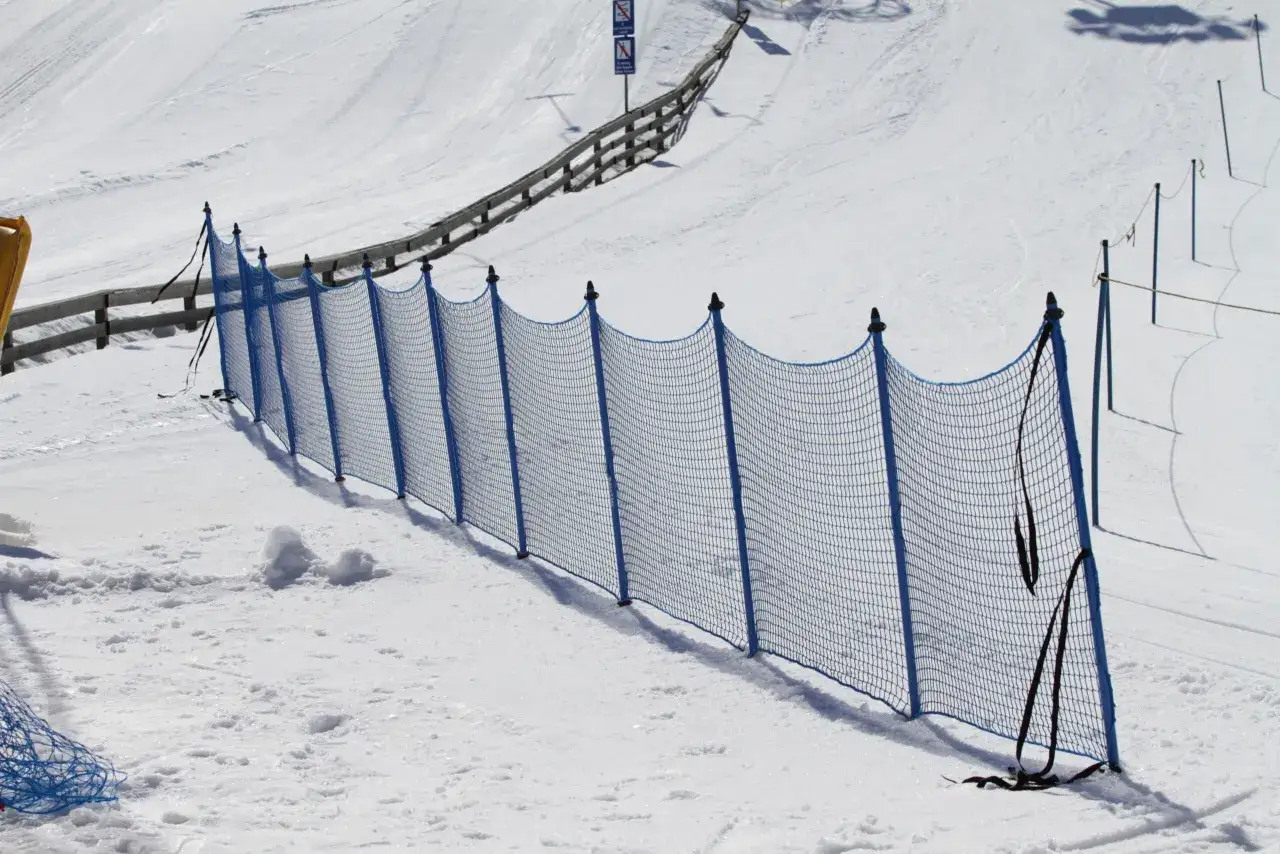 Una valla azul protege la pista de **esquí alpino**. La nieve cubre la ladera, con huellas de esquí visibles.