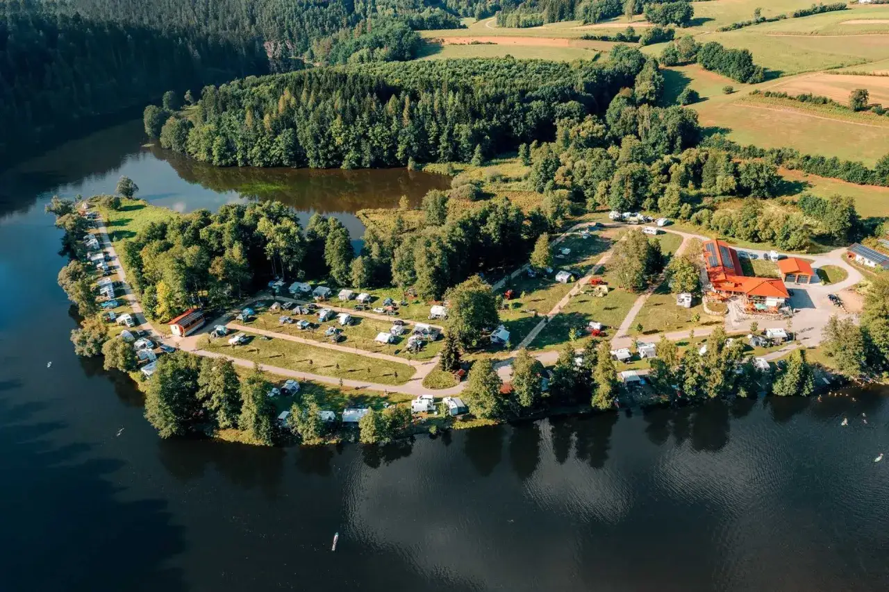Idyllischer bayerischer Wald See mit Campingplatz, Wohnmobilen und Zelten am Ufer. Grüne Wälder und Felder umgeben das glitzernde Wasser.