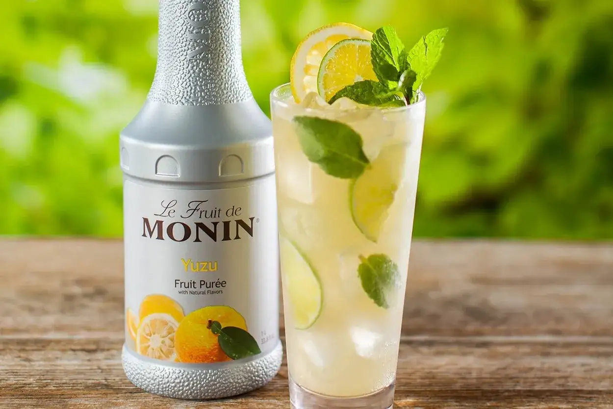 Bouteille de sirop de yuzu Monin et un verre de boisson rafraîchissante avec des tranches de citron, de lime et de la menthe. Parfait pour une recette de sirop de yuzu maison.