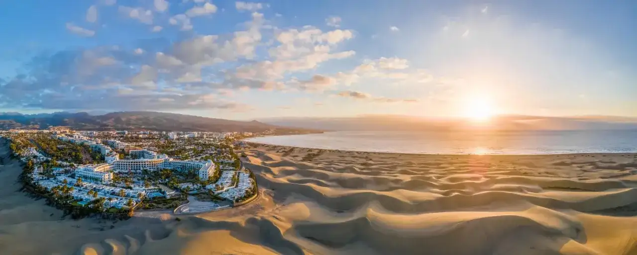 Dunas doradas y resorts blancos junto al mar, un paisaje id&iacute;lico de las mejores playas de Gran Canaria al atardecer.