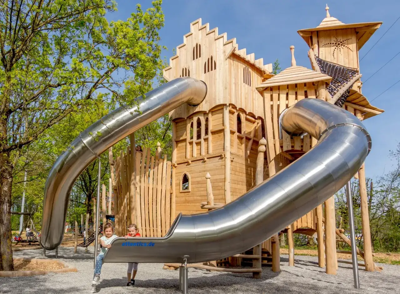 Zwei M&auml;dchen stehen am Fu&szlig;e einer gro&szlig;en Holzburg mit Rutschen. Ein perfekter Ort f&uuml;r eine **Camping Familie** voller Abenteuer.