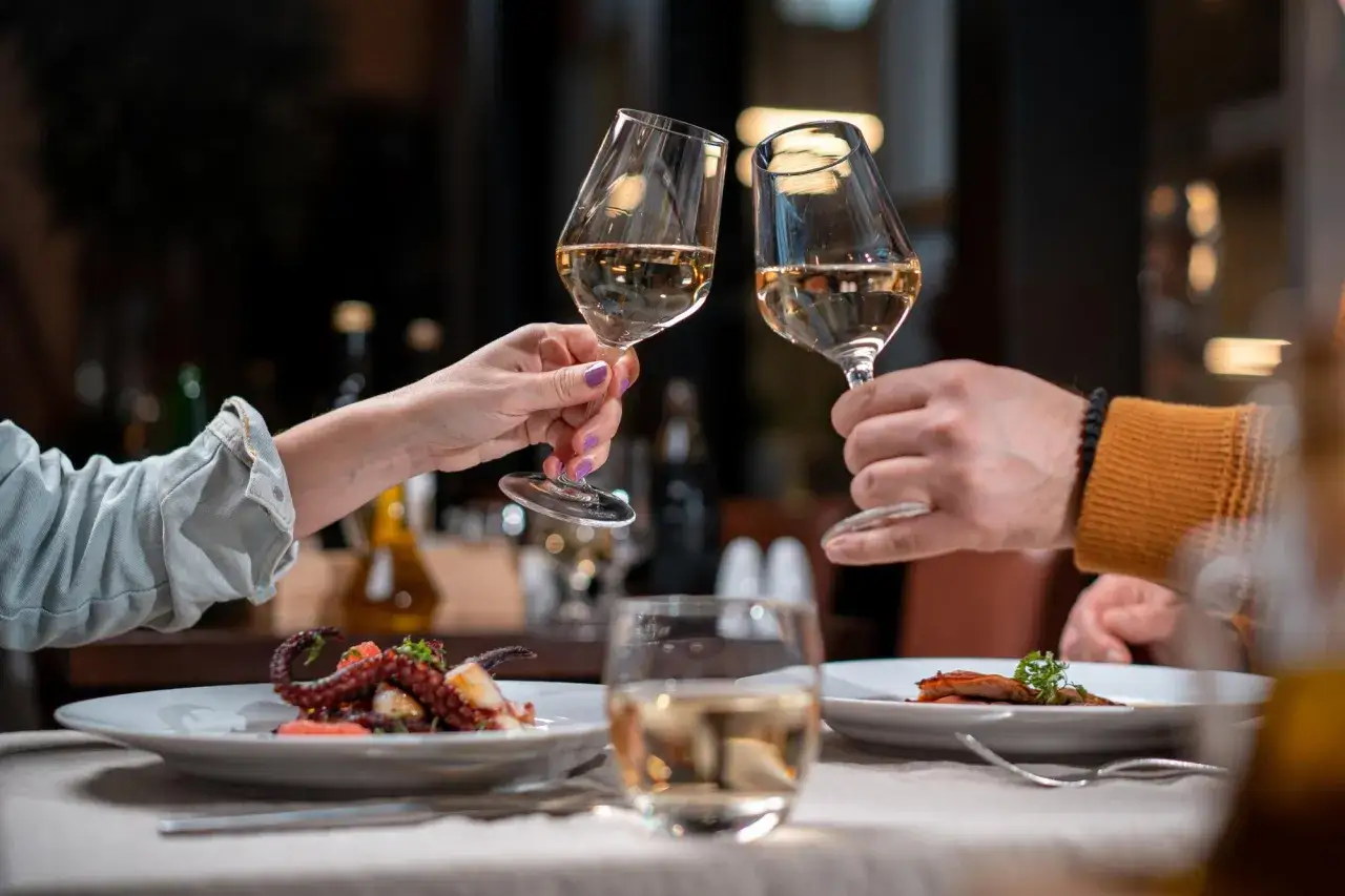 Dwie ręce wznoszą kieliszki białego wina do toastu. Na talerzach dania, a w tle atmosfera sprzyjająca romantycznym chwilom i polskim piosenkom na rocznicę ślubu.