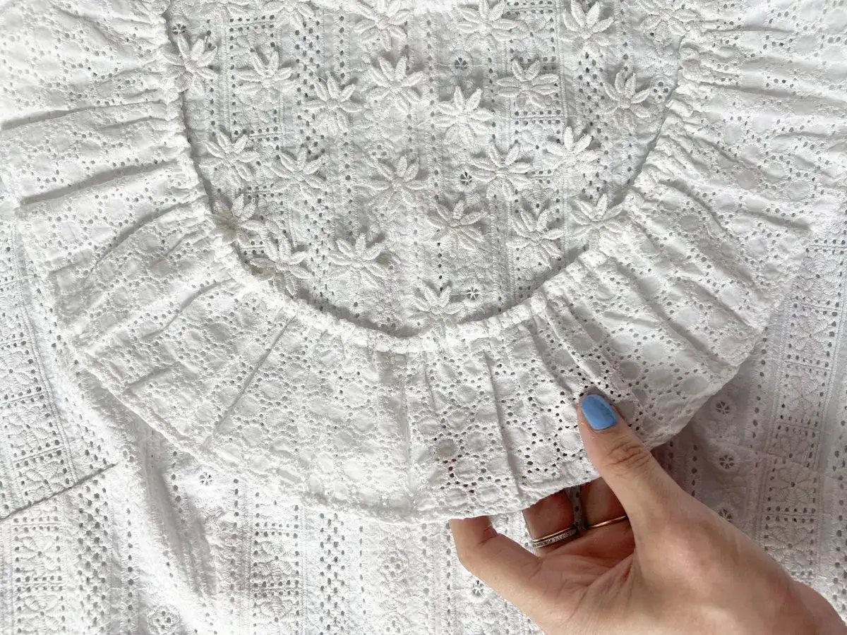 Close-up of a white eyelet fabric, showing intricate floral embroidery. A hand gently holds the fabric, hinting at how to make a lace collar.