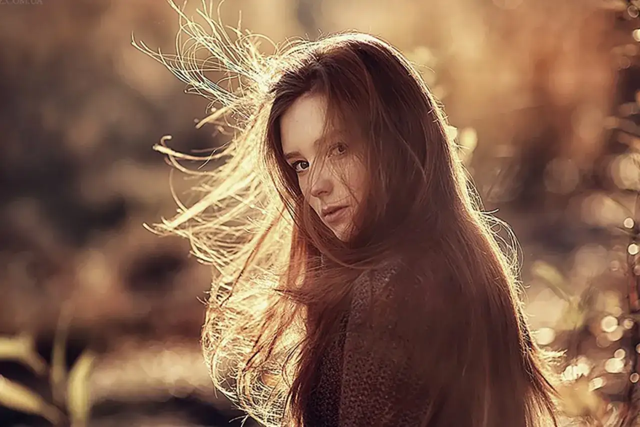 Jeune femme aux cheveux roux, éclairée par un soleil bas. Sa chevelure s'envole dans un souffle d'air, créant une **photo contre jour** poétique.