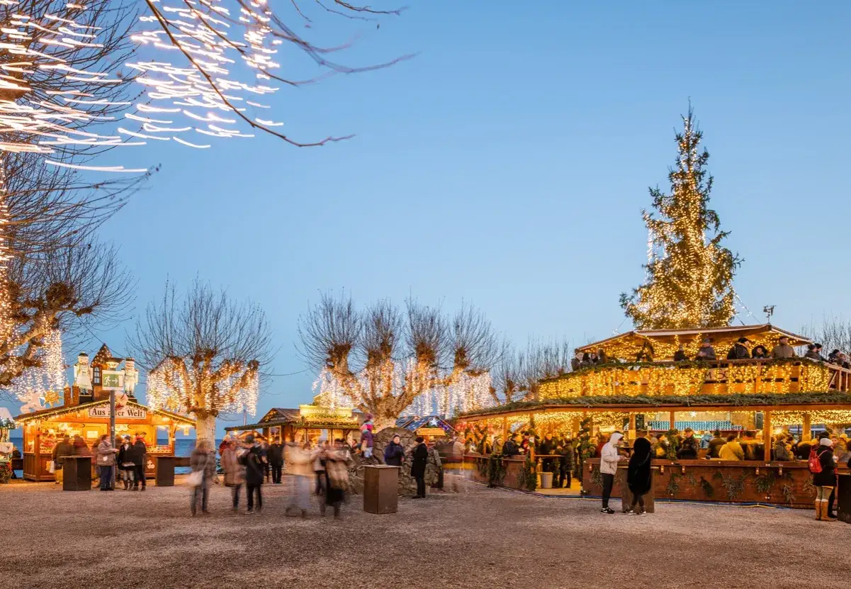 Der **Weihnachtsmarkt Konstanz** erstrahlt in festlichem Glanz. Menschen flanieren zwischen beleuchteten Buden und einem gro&szlig;en Weihnachtsbaum.