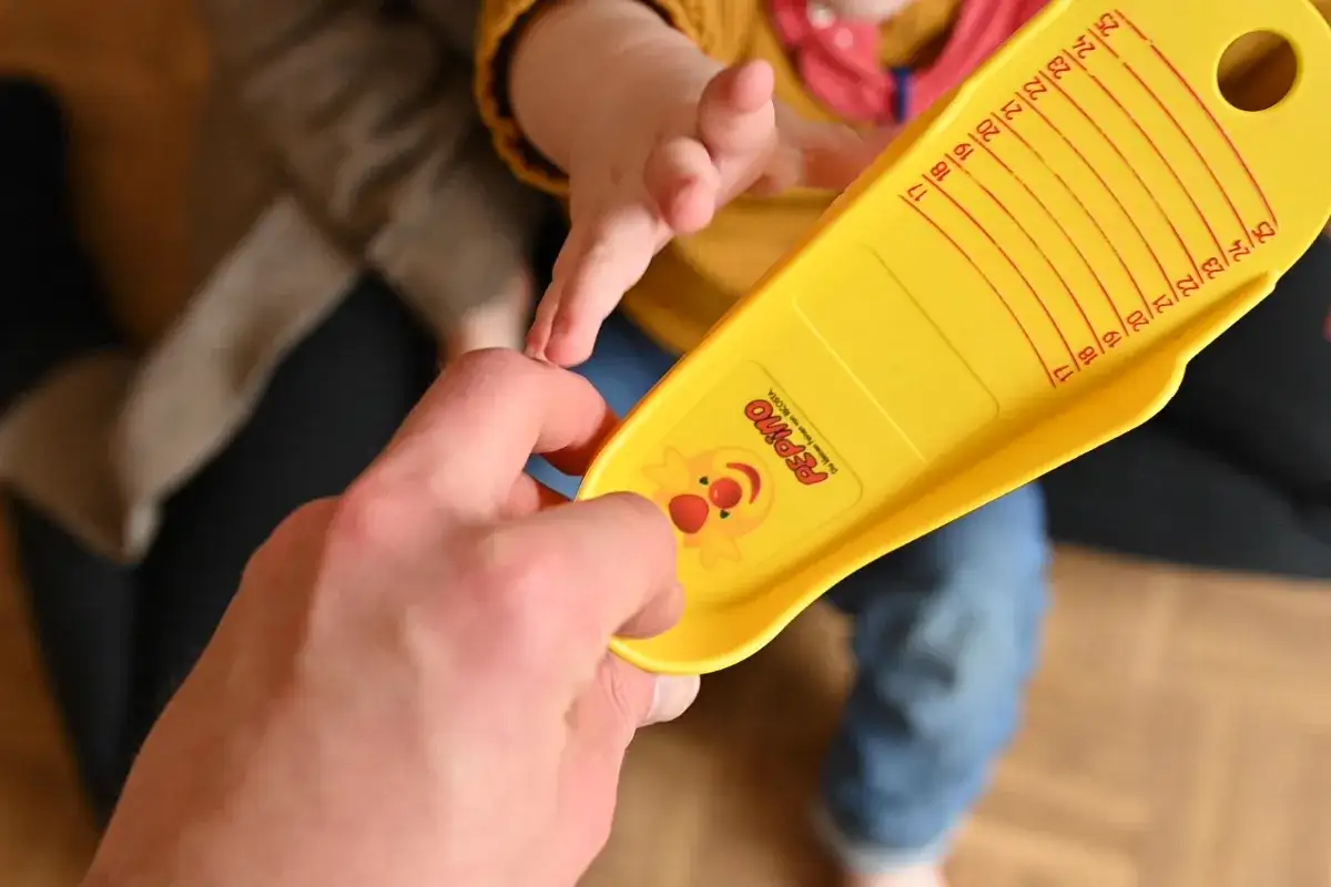 Ein gelber Fußmesser zeigt, wie fallen Trollkids Schuhe aus. Eine Hand hält ihn, ein Babyfuß wird gemessen.