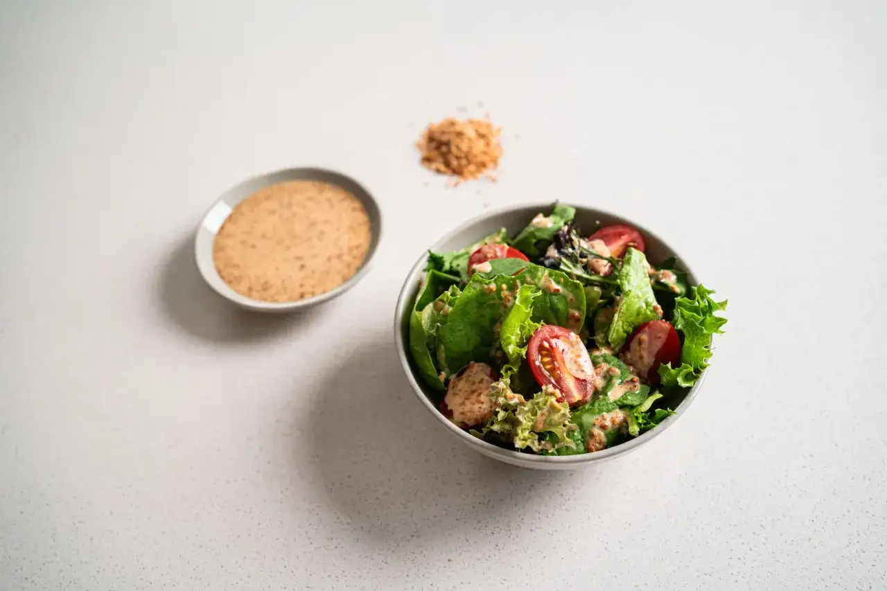 A fresh salad with tomatoes and lettuce is drizzled with a creamy roasted sesame dressing. A small dish of the dressing sits nearby.