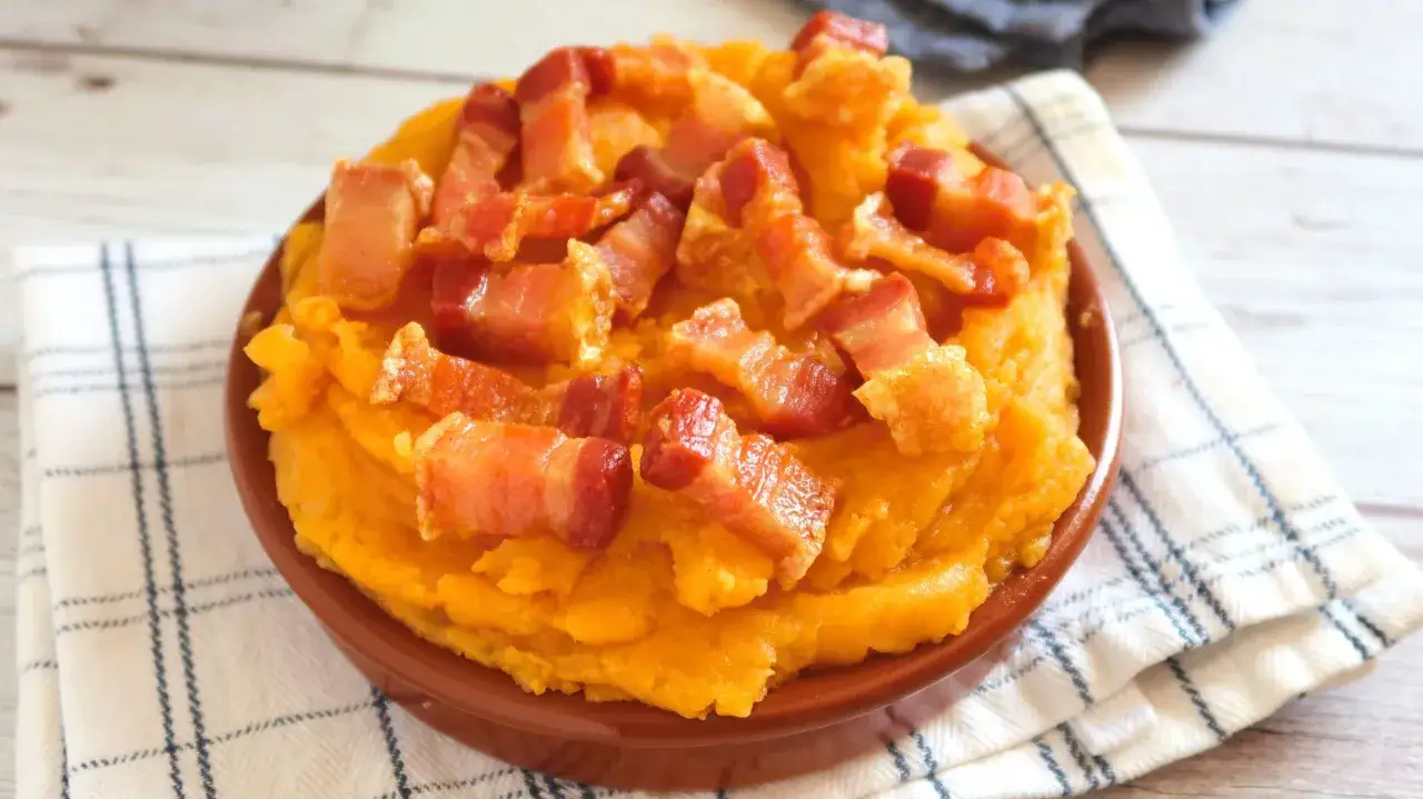 Patatas revolconas cremosas con trozos de bacon crujiente, servidas en un cuenco de barro sobre un paño a cuadros.