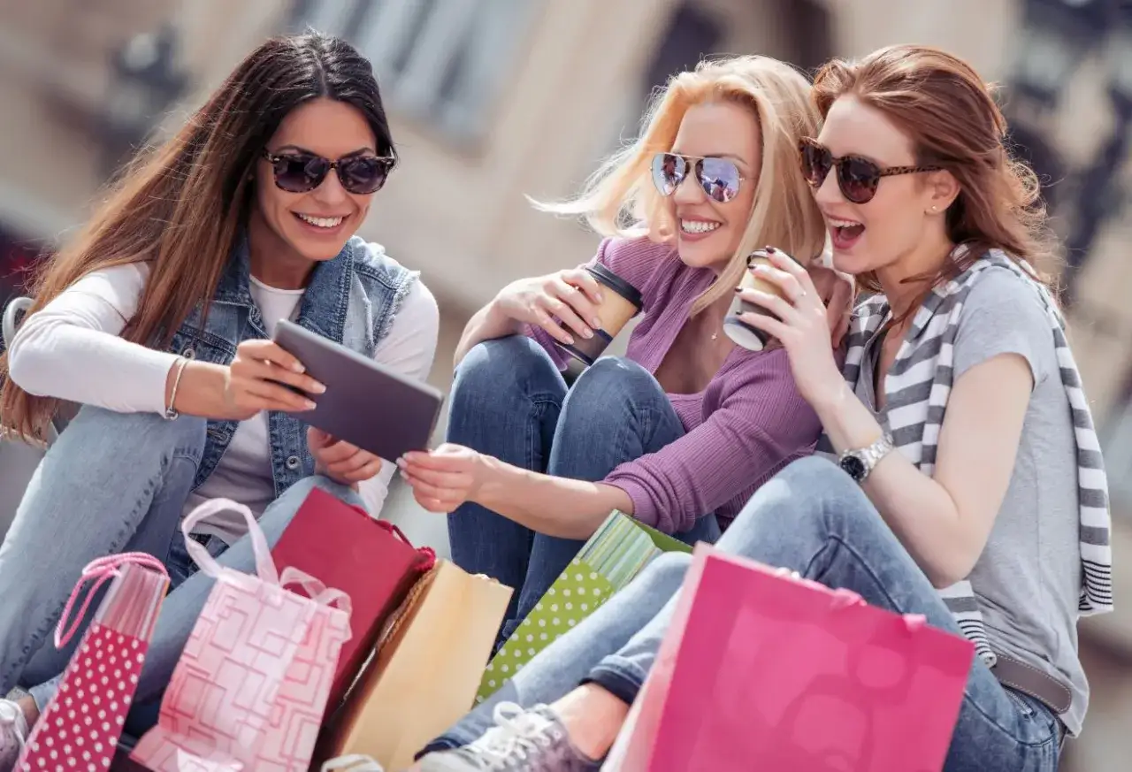Trzy przyjaciółki cieszą się wspólnym live shoppingiem, przeglądając tablet wśród kolorowych torebek z zakupami.