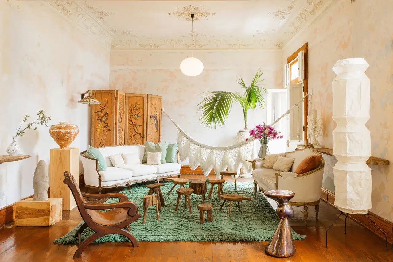Przytulny salon w stylu boho z białymi sofami, makramowym hamakiem, drewnianymi stołkami i egzotycznymi roślinami.