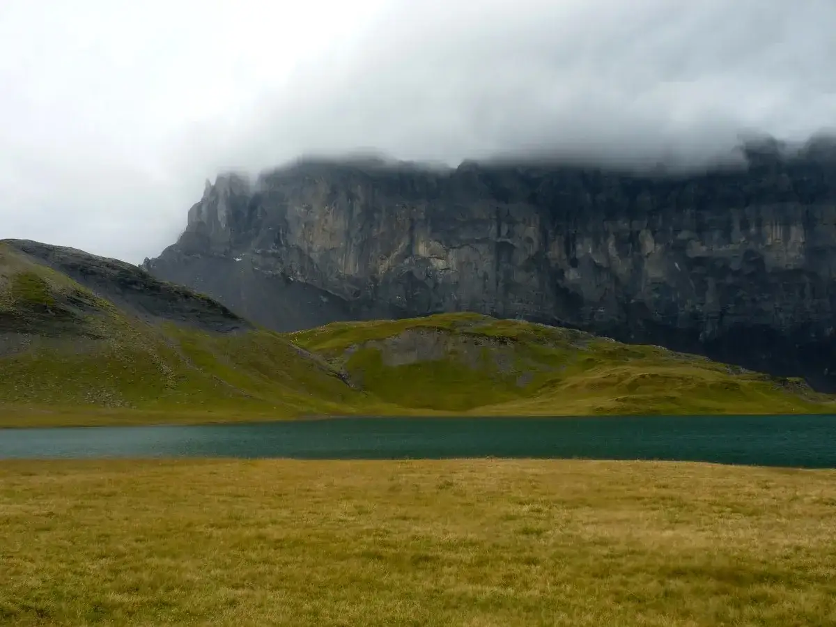 Vaste paysage alpin avec un lac turquoise, des collines verdoyantes et une imposante paroi rocheuse voilée de nuages, évoquant le **tour des Fiz**.
