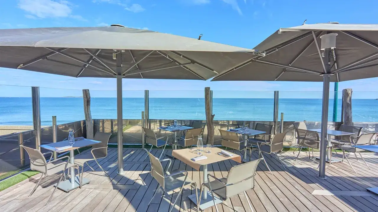 Terrasse d'un bistro &agrave; Saint-Malo avec vue sur la mer. Tables dress&eacute;es sous de grands parasols, ambiance estivale.