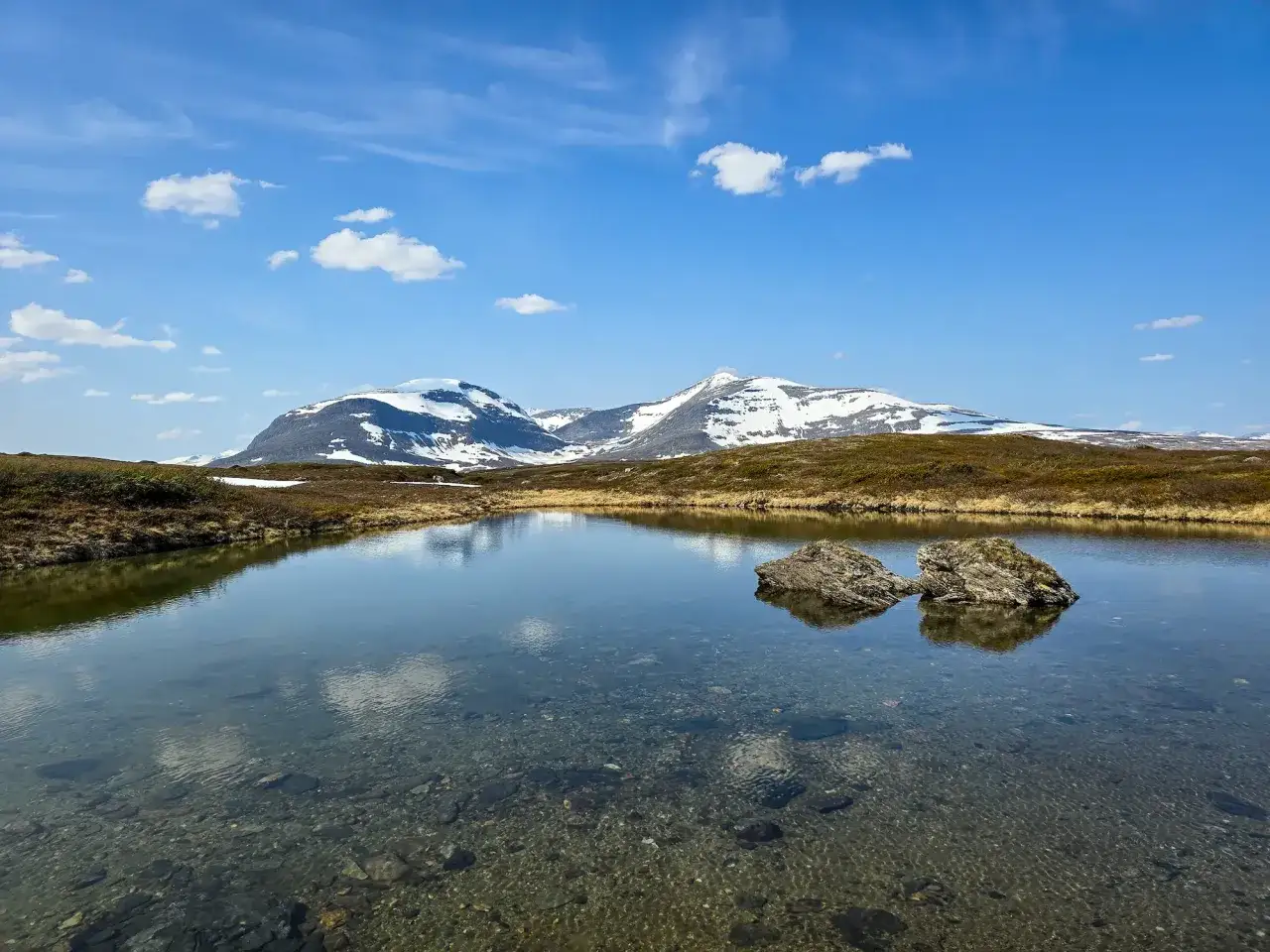 En klarblå himmel med moln speglar sig i en lugn sjö. Snöklädda berg reser sig i bakgrunden, en idyllisk vy för vandring i Ramundberget.