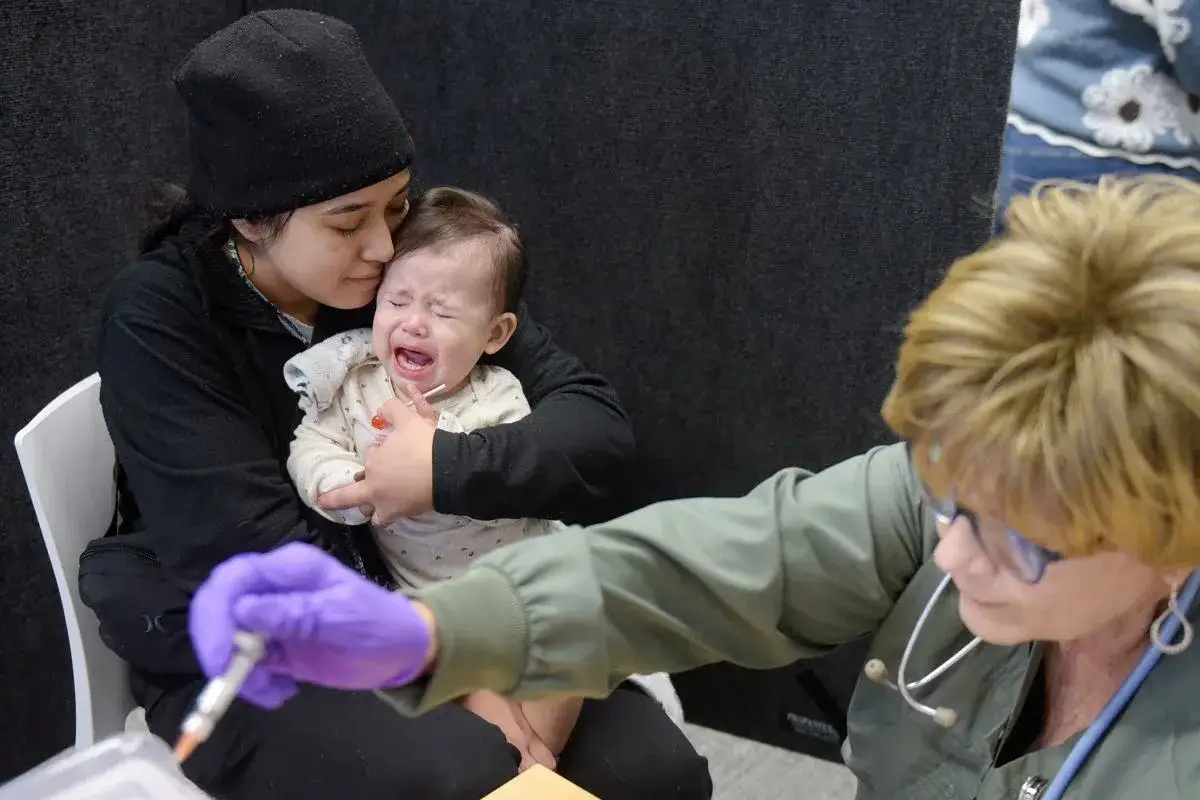 Mama tuli dziecko, które płacze przed szczepieniem MMR. Pielęgniarka w fioletowej rękawiczce przygotowuje zastrzyk.