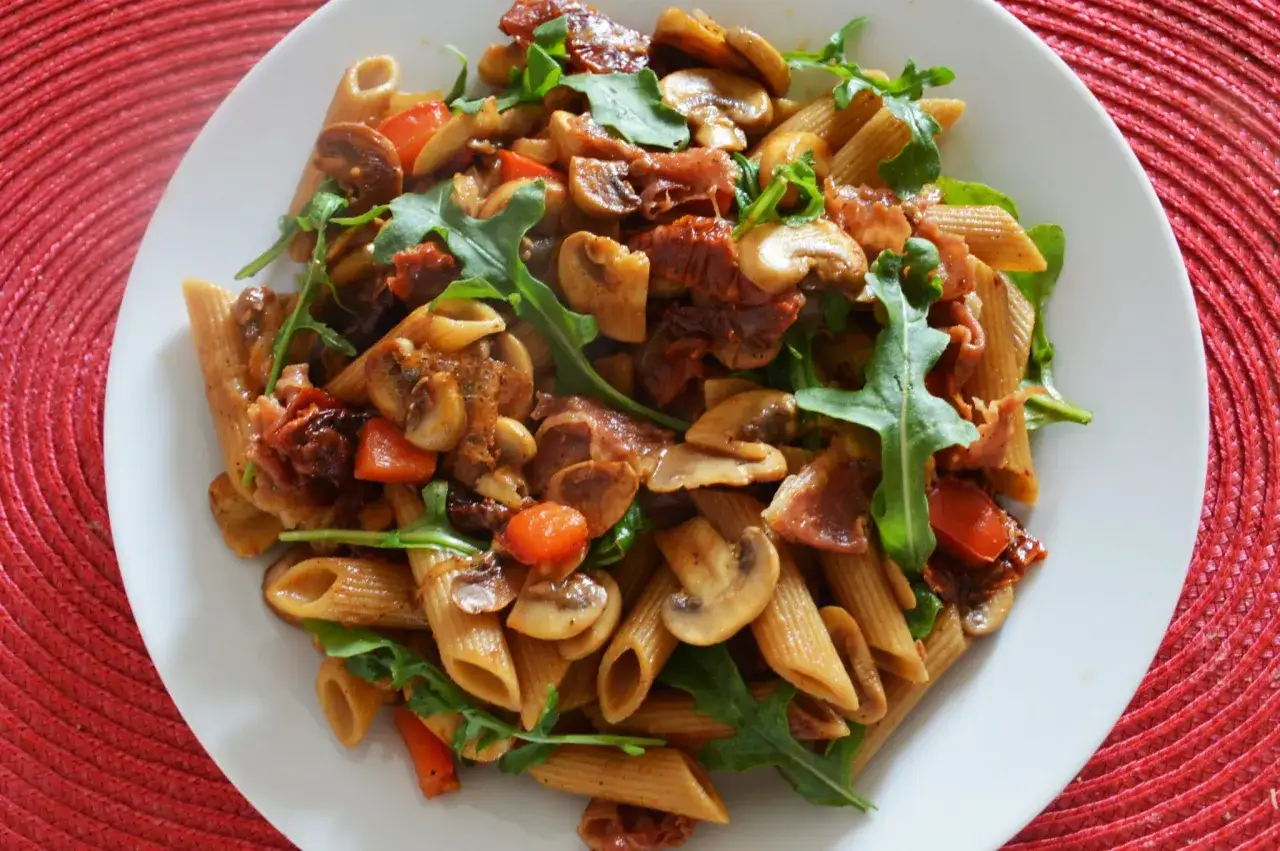 Penne z szynką parmeńską, pieczarkami, pomidorkami suszonymi i rukolą. Pyszne danie na talerzu.