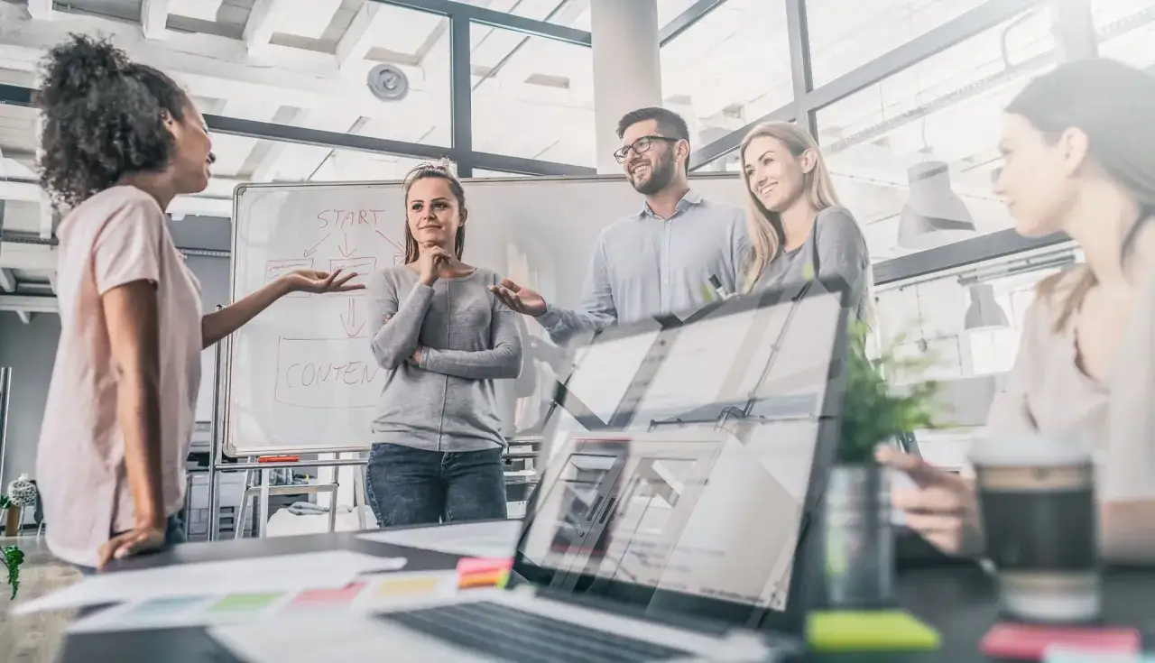 Team bespricht Strategie f&uuml;r Ausbildung Marketingkommunikation. Eine Frau erkl&auml;rt am Whiteboard, andere h&ouml;ren zu.