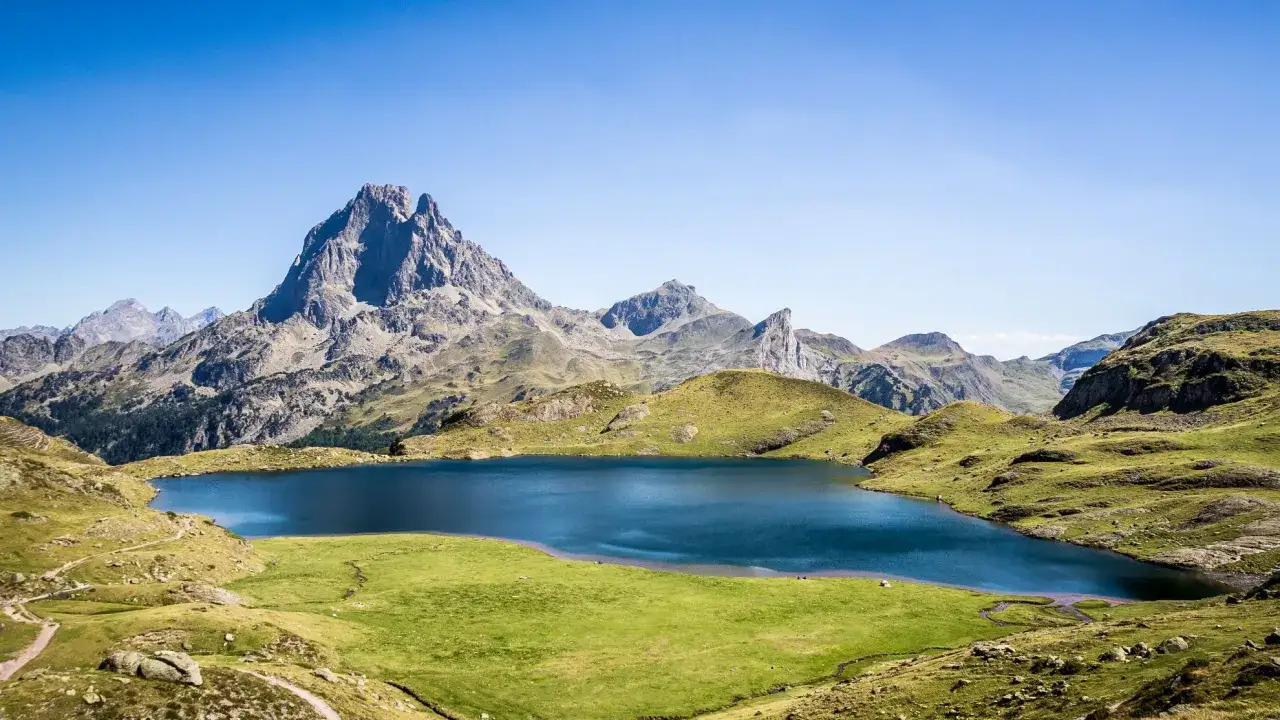 Lago azul sereno en el **Valle de Ossau**, rodeado de verdes colinas y picos montañosos imponentes bajo un cielo despejado.