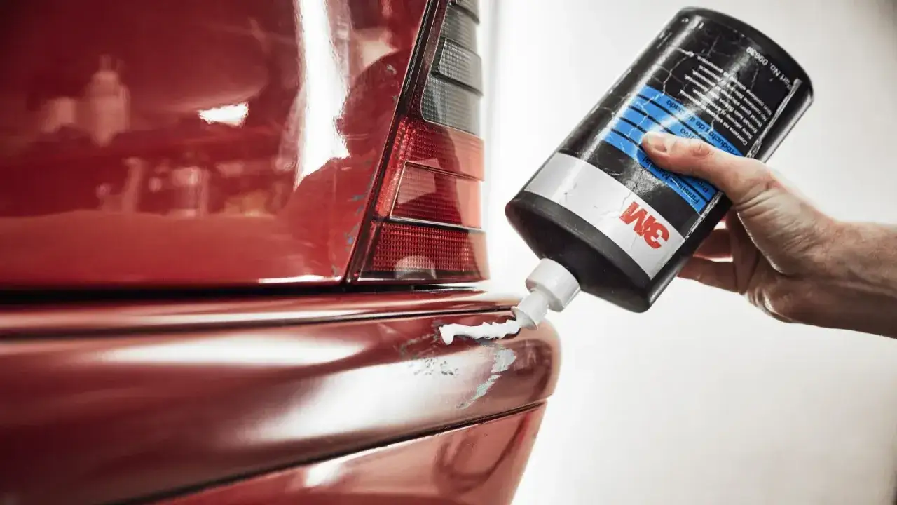 Roter Autolack mit tiefen Kratzern bis auf Grundierung. Eine Hand hält eine 3M-Flasche mit Polierpaste.