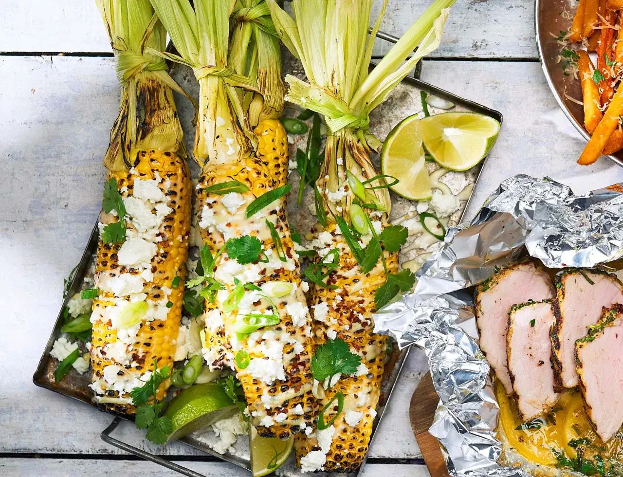 Grillade majskolvar, corn ribs, toppade med ost, örter och lime. Bredvid ligger skivat fläskkött i folie.