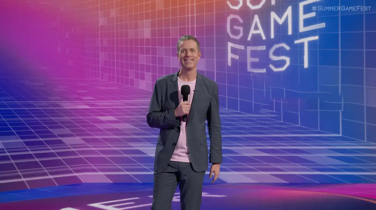Uśmiechnięty prowadzący z mikrofonem na scenie z napisem #SUMMERGAMEFEST.
