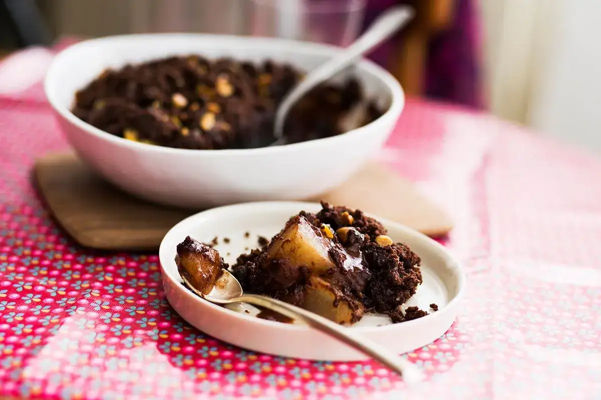 Un délicieux crumble chocolat avec des morceaux de poire fondants, servi dans une assiette blanche sur une nappe rose à motifs.