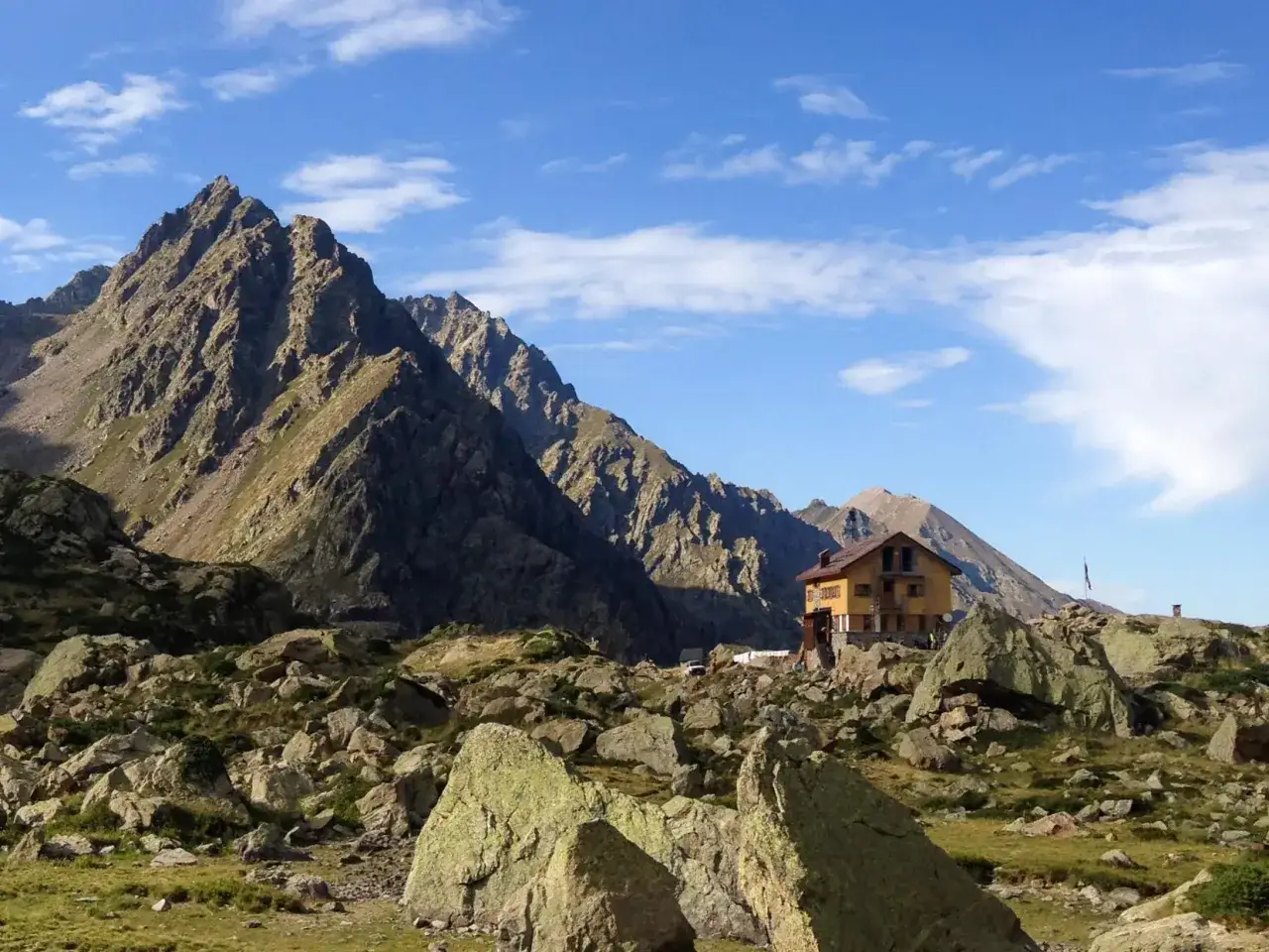 Il rifugio Genova, incastonato tra imponenti montagne rocciose e un cielo azzurro con nuvole bianche, offre un riparo solitario in un paesaggio alpino mozzafiato.
