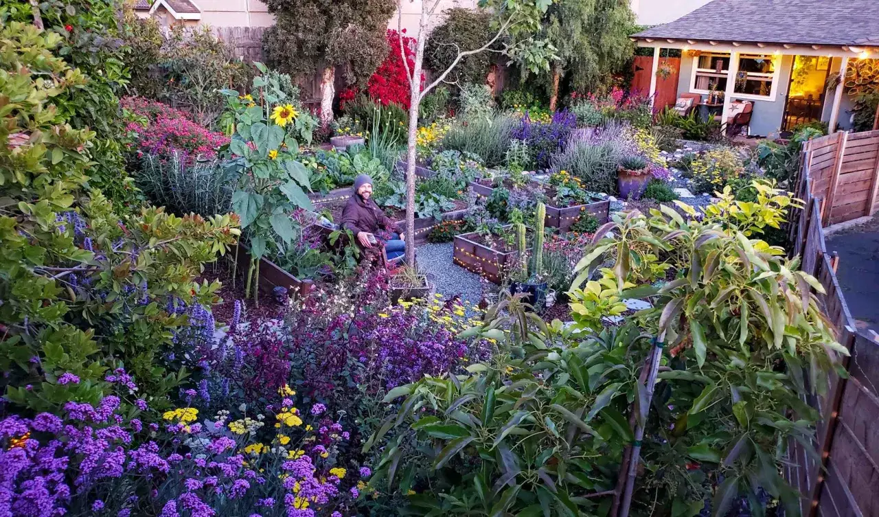 Ein Mann sitzt in einem üppigen, tierfreundlichen Garten voller bunter Blumen und Gemüsebeete.