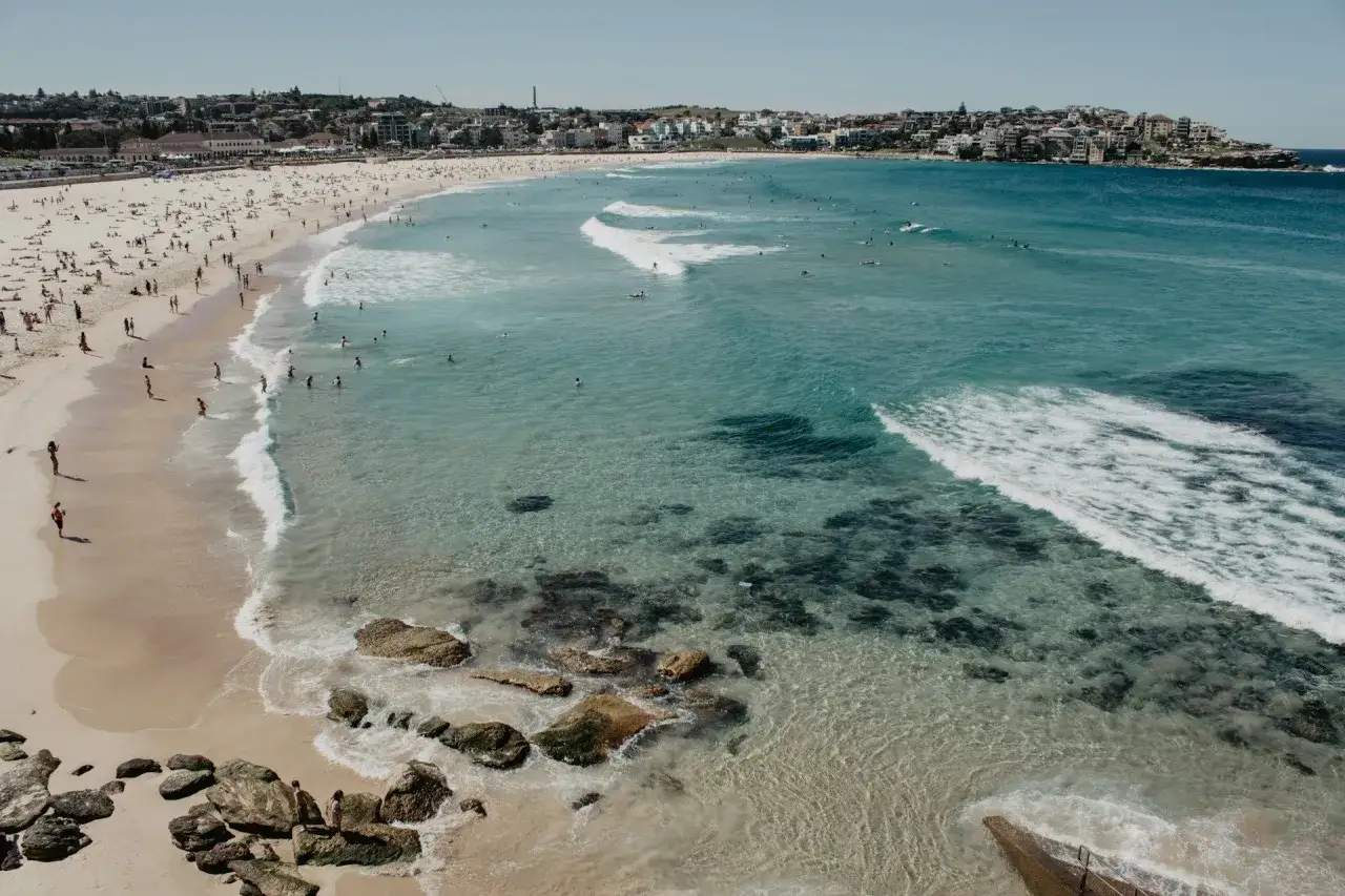 Słoneczne lato w Australii. Tłumy ludzi na piaszczystej plaży i w turkusowej wodzie, surferzy łapią fale.
