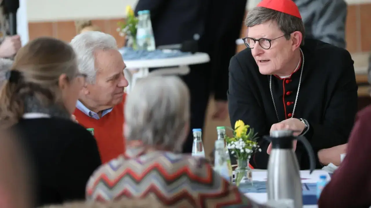 Kardinal im Gespräch, während eine Frau mit einem bunten Pullover und ein Mann im orangen Pullover zuhören. Möglicherweise eine Diskussion über Kritik an der Kirche.