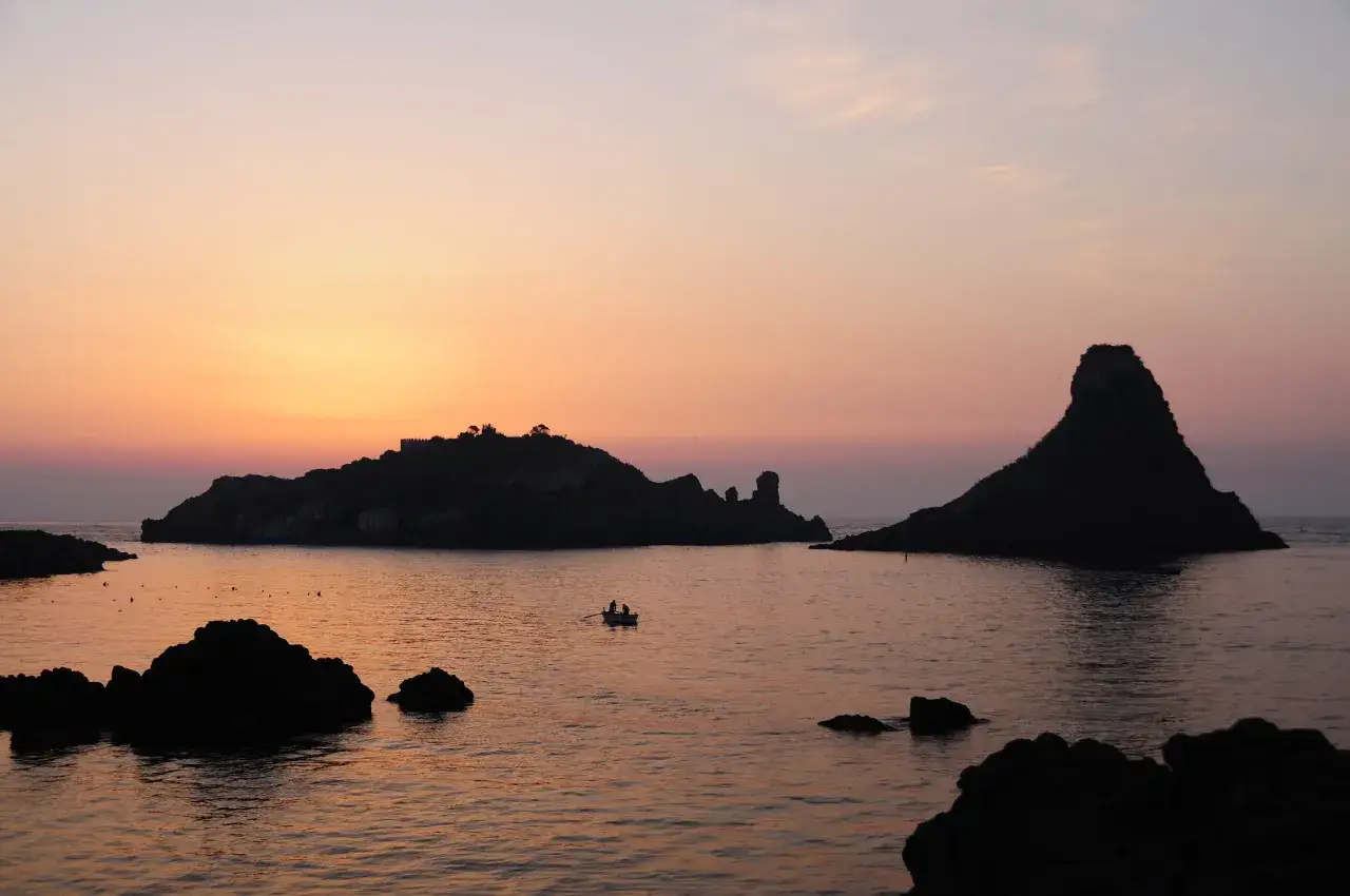 Tramonto ad Aci Trezza, con i faraglioni che emergono dal mare calmo. Una barca con due pescatori solca le acque rosate.