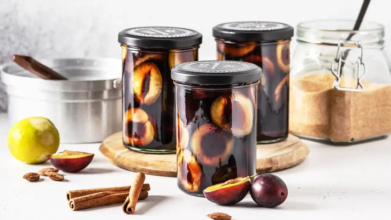 Drei Gl&auml;ser mit eingelegten Pflaumen, bereit zum Einkochen. Daneben liegen frische Pflaumen, Zimtstangen, Mandeln und eine Zitrone.