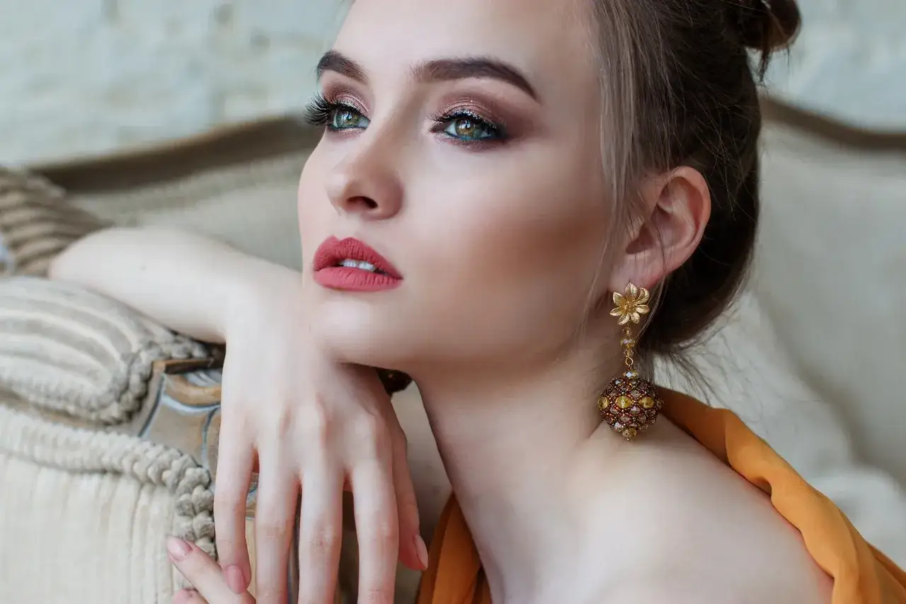Mujer con piel clara, ojos verdes y labios rojos, luciendo un elegante pendiente dorado.