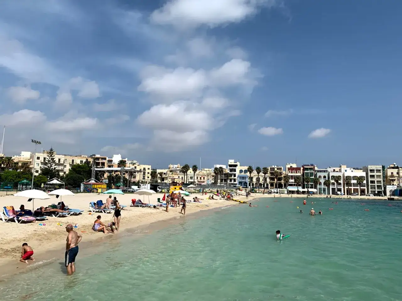 Słoneczny dzień na **pretty bay**. Ludzie odpoczywają na plaży, kąpią się w krystalicznie czystej wodzie, a w tle widać kolorowe budynki.