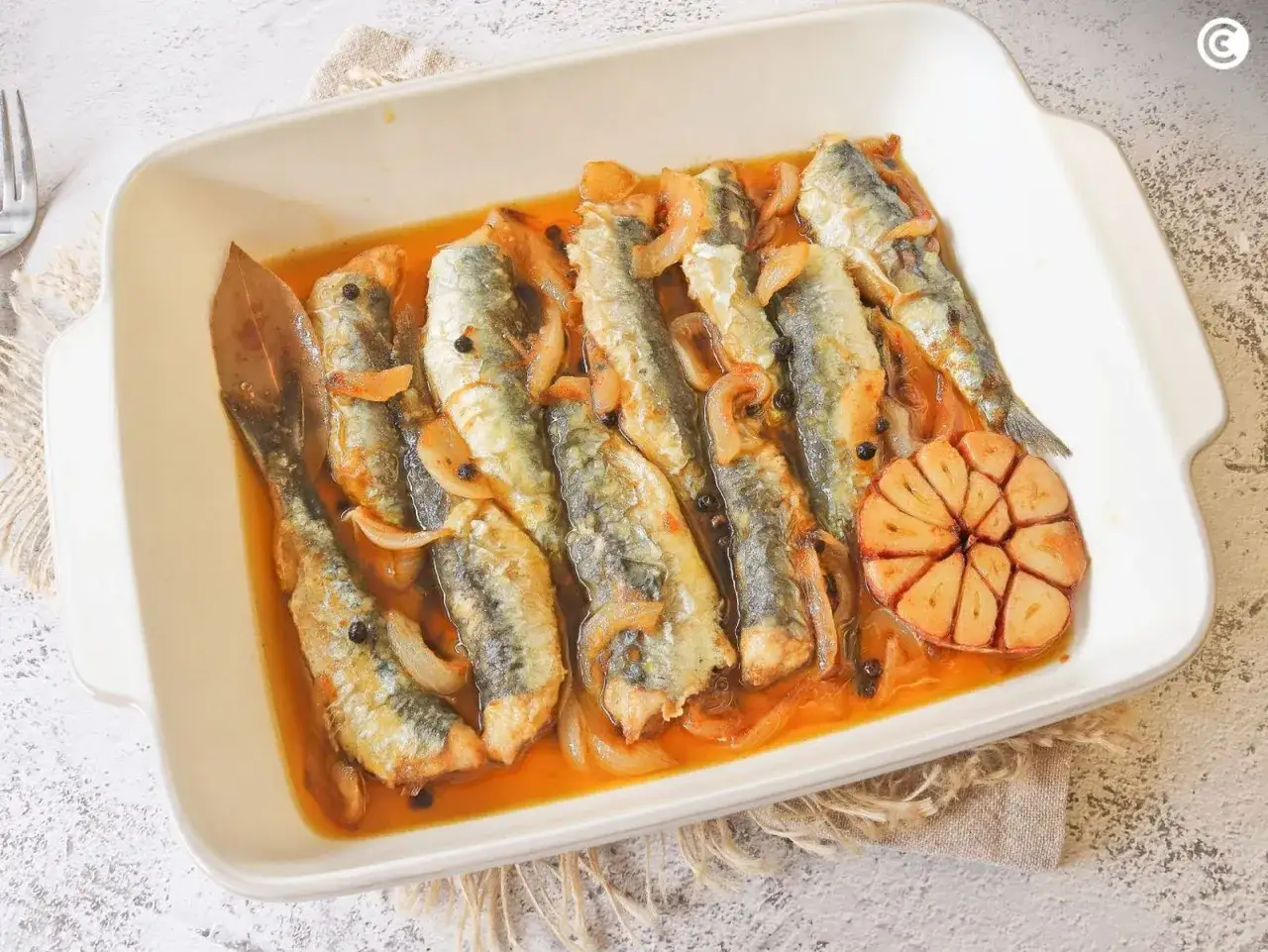 Sardinas en escabeche con cebolla, granos de pimienta y una rodaja de ajo asado en una fuente blanca.