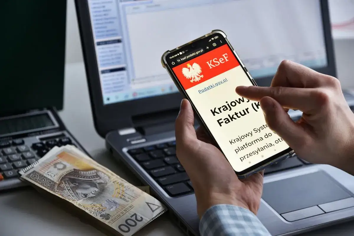 Dłoń trzyma smartfon z aplikacją KSeF, obok leżą pieniądze i kalkulator. Czy wonga dzwoni do pracodawcy?