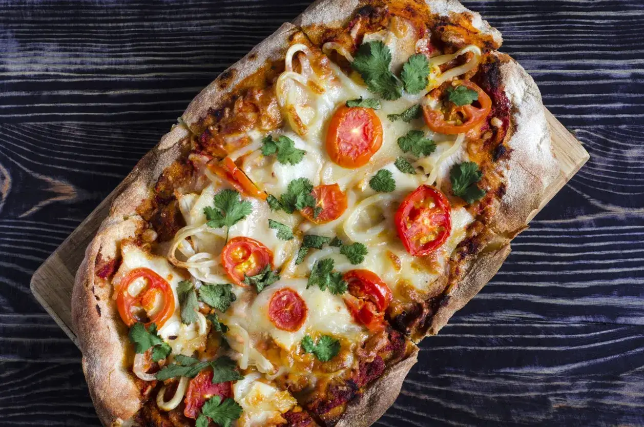 En avlång romersk pizza med tomater, lök och färska örter, serverad på en träplatta över en mörk träbakgrund.