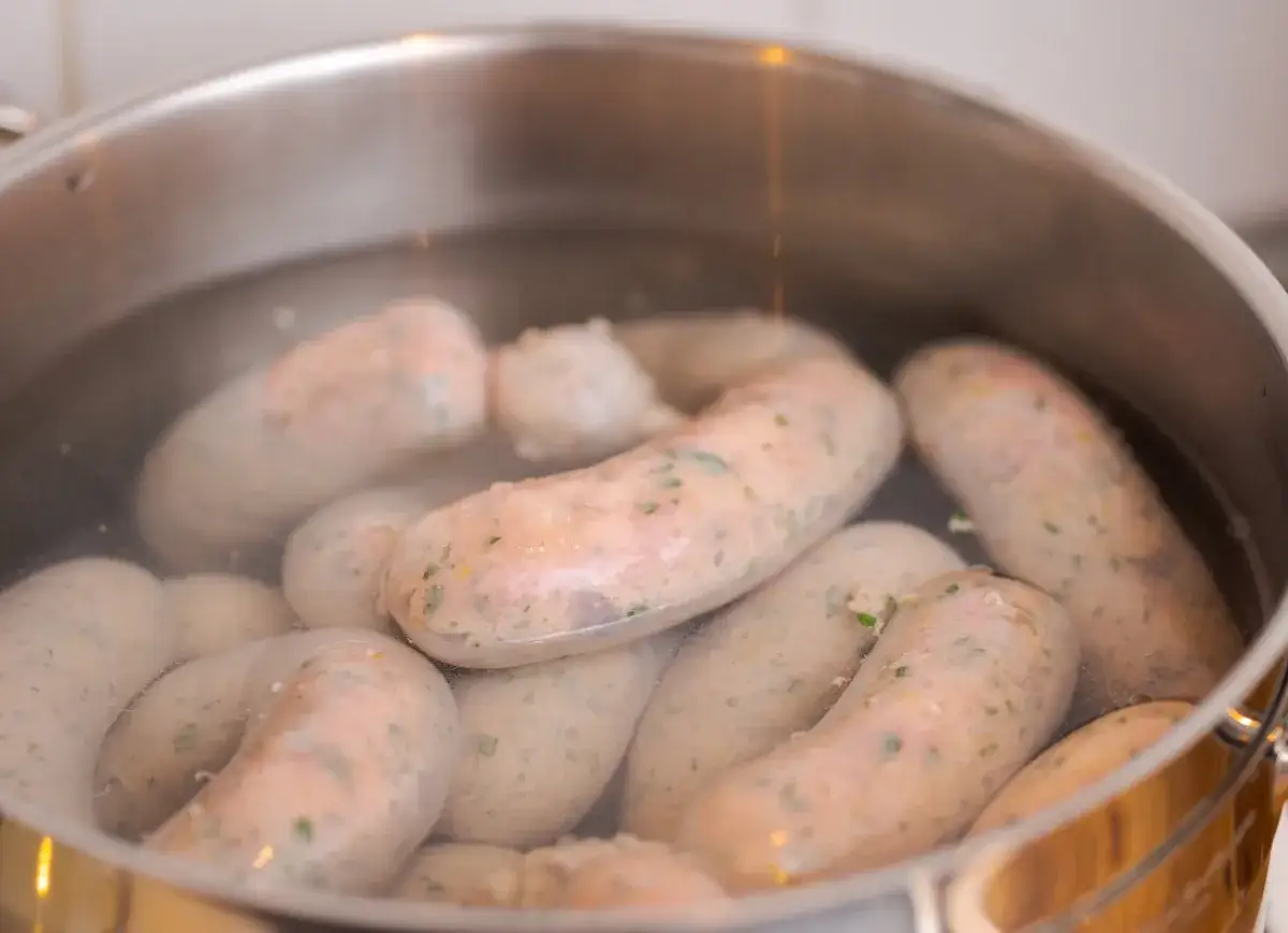 Kiełbasa biała parzona gotuje się w garnku. Widać wiele grubych kiełbasek w wodzie, niektóre z zielonymi ziołami.