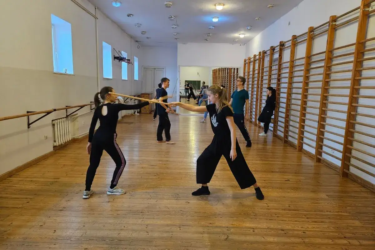 Dwie studentki ćwiczą walkę kijami na WF-ie. W tle inni studenci i drabinki gimnastyczne.