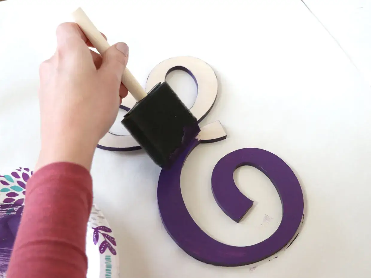 Maintenant, comment décorer une lettre ! Une main peint une lettre en bois en violet avec un pinceau éponge.