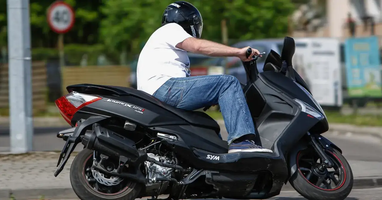 Mężczyzna w kasku jedzie na skuterze SYM Joymax 125. Znak ograniczenia prędkości do 40 km/h sugeruje, że nie osiąga on swojej maksymalnej prędkości.