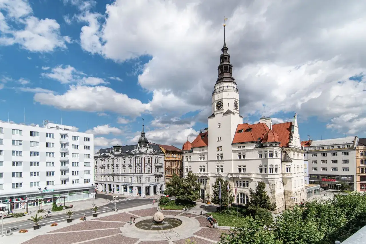 Ein sonniger Tag in Opava, Tschechien. Ein Marktplatz mit Brunnen und historischen Geb&auml;uden, darunter ein Rathausturm mit Uhr.