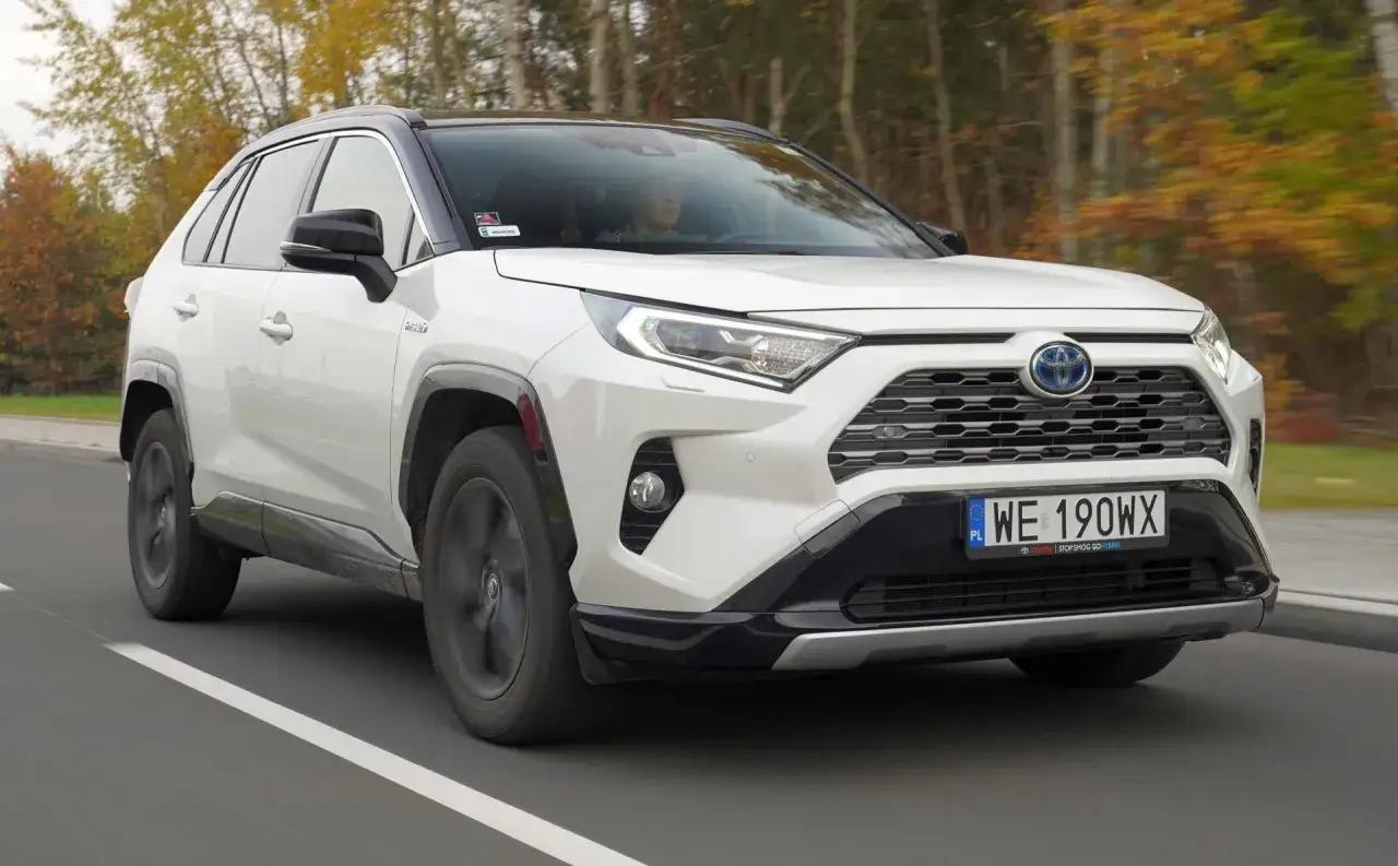 Biały Toyota RAV4 2022 z czarnym dachem jedzie po drodze, otoczony jesiennymi drzewami.