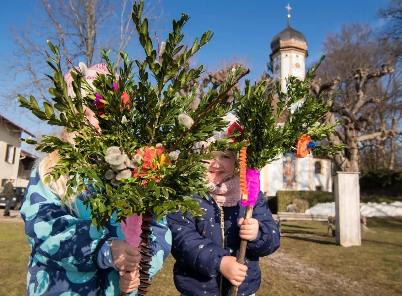 Kinder halten geschmückte Palmbuschen vor einer Kirche. Ein fröhlicher Anblick am Palmsonntag 2026.