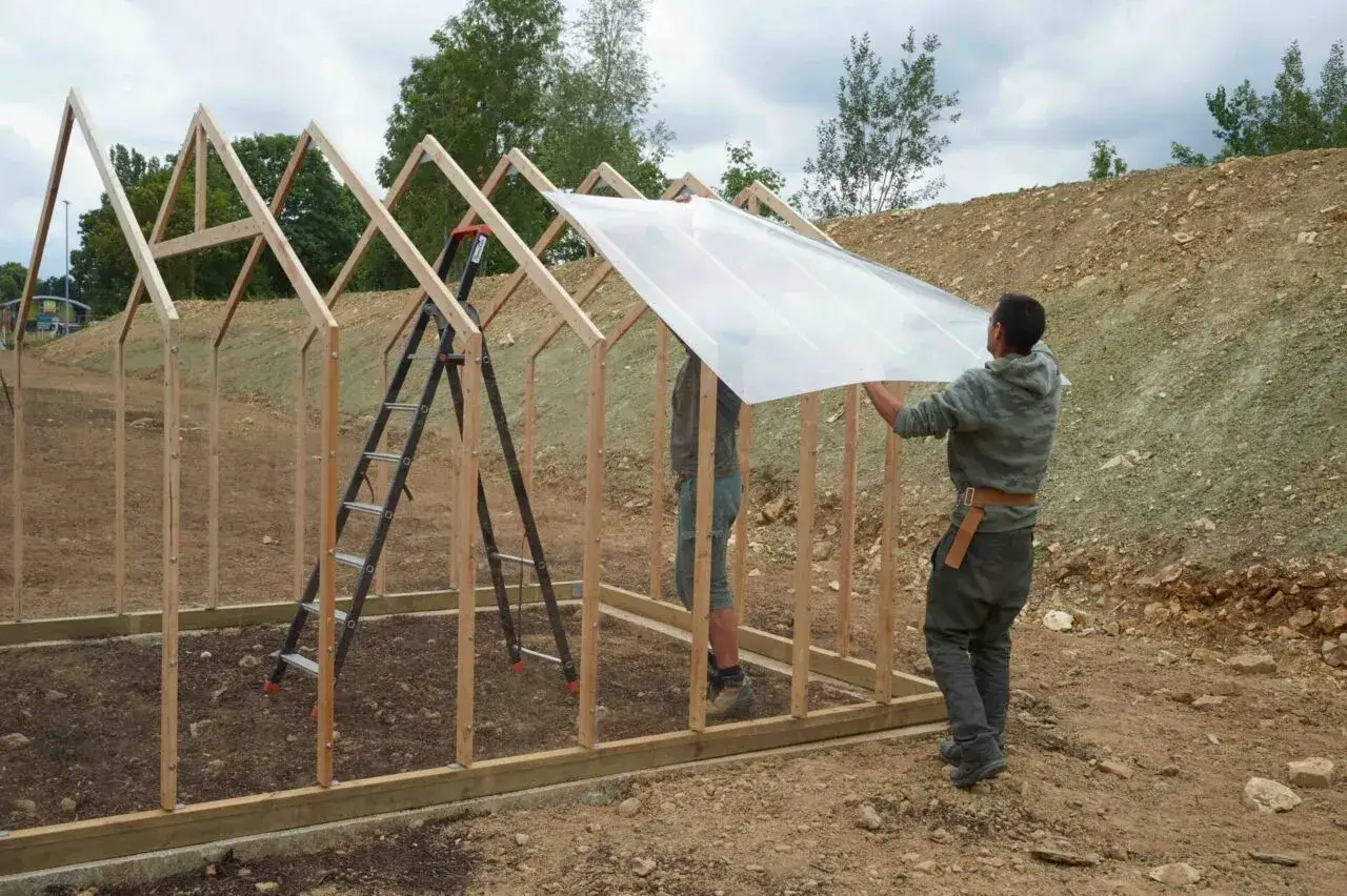 Deux hommes travaillent &agrave; fabriquer une serre en bois, posant une grande plaque de plastique transparent sur la structure.