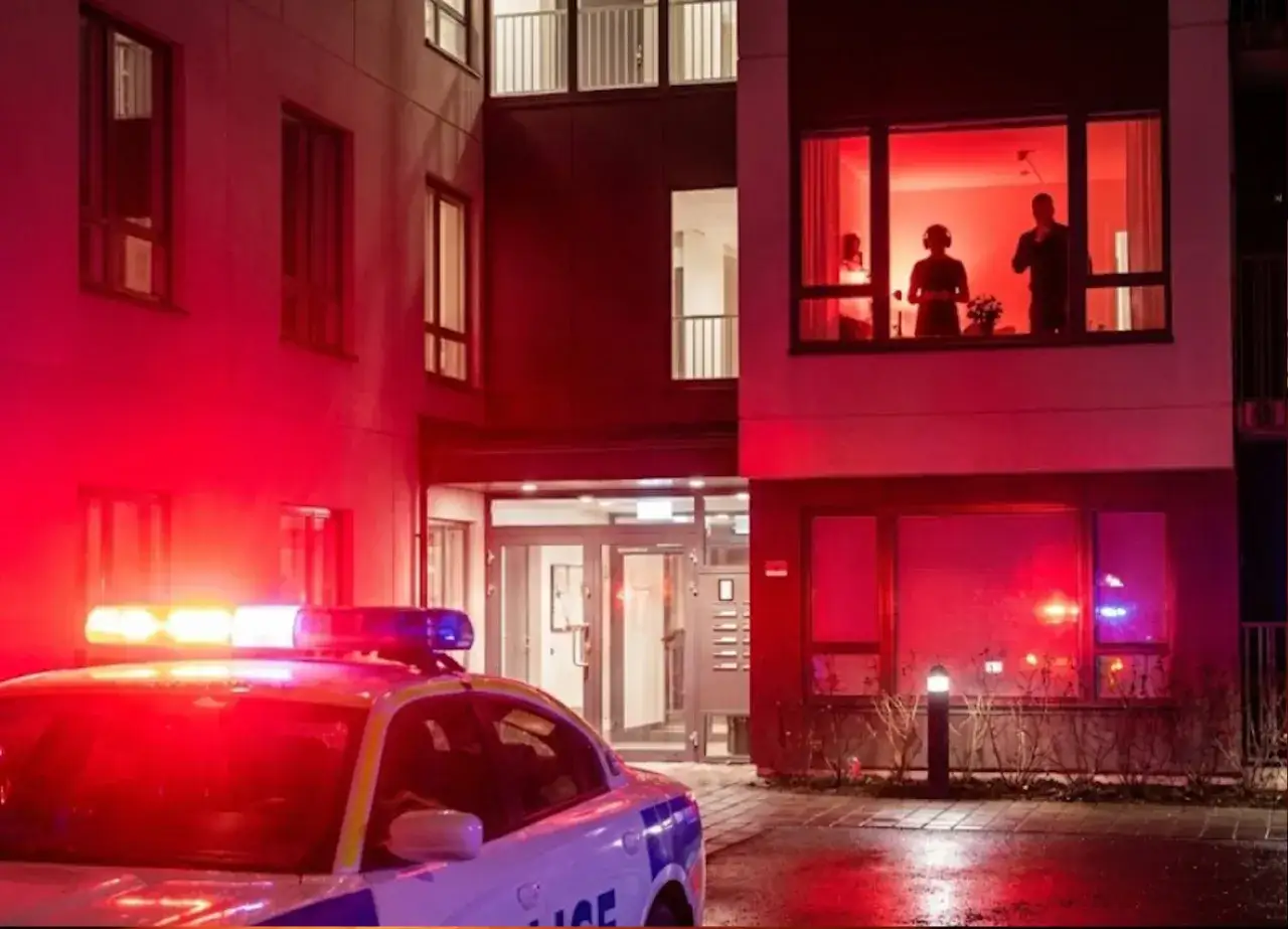 Radiowóz policyjny z włączonymi sygnałami świetlnymi przed budynkiem, gdzie w oknie widać sylwetki ludzi. Może to być interwencja związana z zakłóceniem ciszy nocnej.