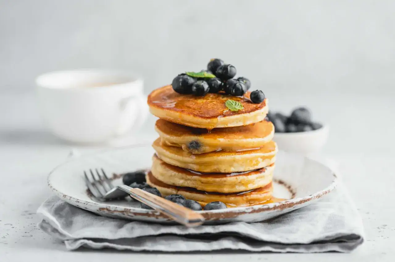 Pila di pancake con farina di riso, guarniti con mirtilli freschi e sciroppo d'acero. Una tazza di caff&egrave; sullo sfondo.